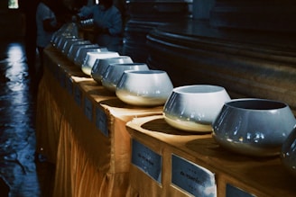 A row of shiny metallic bowls resting on a covered table, with soft lighting creating reflections on their surfaces. The background is dimly lit, with indistinct figures engaging with the setup, possibly participating in a ritual or event.