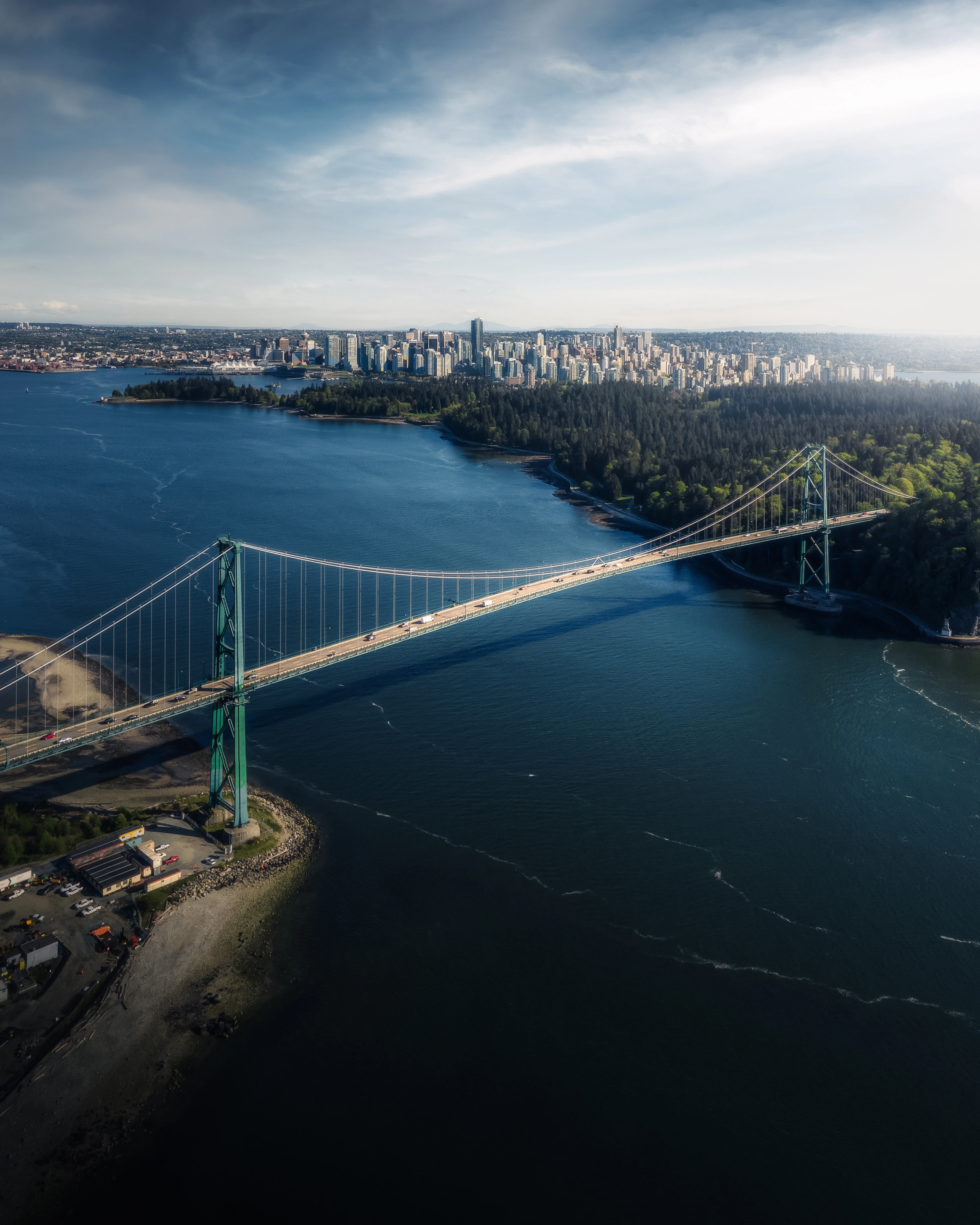Aerial view of the Lions Gate Bridge spanning the water toward downtown Vancouver's skyline under a blue sky.
