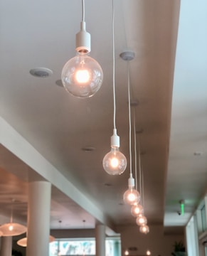 A series of large, clear light bulbs hang in a straight line from the ceiling, each lit with a warm glow. The bulbs are suspended by long white wires in a minimalist interior space featuring cream-colored columns and soft neutral tones.