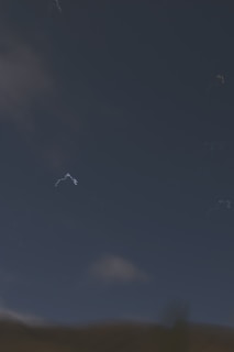 A dark sky occupies the upper half of the image with scattered, faint cloud formations. The lower part of the image shows a dark, indistinct landscape that transitions from a dark blue-green to a muted brown. Subtle scattered white streaks appear sporadically against the night sky, hinting at perhaps faint stars or optical artifacts.