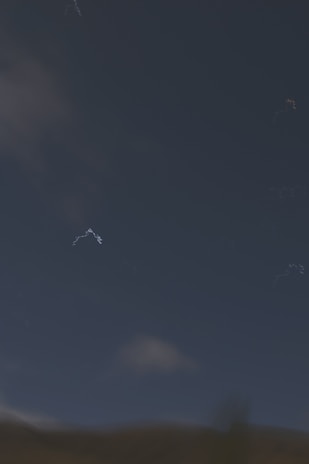 A dark sky occupies the upper half of the image with scattered, faint cloud formations. The lower part of the image shows a dark, indistinct landscape that transitions from a dark blue-green to a muted brown. Subtle scattered white streaks appear sporadically against the night sky, hinting at perhaps faint stars or optical artifacts.
