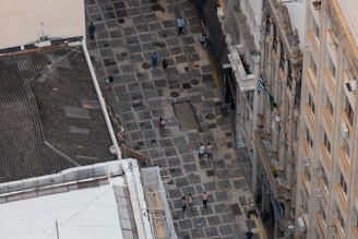 Aerial view of a stone-paved pedestrian street lined with historic buildings. Several people are walking along the street, which is flanked by buildings with ornate facades and balconies. Brazilian flags are visible hanging from the balconies.