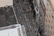 Aerial view of a stone-paved pedestrian street lined with historic buildings. Several people are walking along the street, which is flanked by buildings with ornate facades and balconies. Brazilian flags are visible hanging from the balconies.