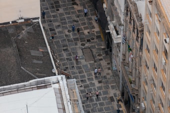 Aerial view of a stone-paved pedestrian street lined with historic buildings. Several people are walking along the street, which is flanked by buildings with ornate facades and balconies. Brazilian flags are visible hanging from the balconies.