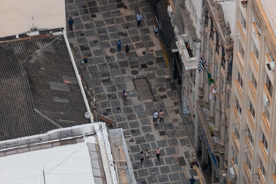 Aerial view of a stone-paved pedestrian street lined with historic buildings. Several people are walking along the street, which is flanked by buildings with ornate facades and balconies. Brazilian flags are visible hanging from the balconies.