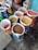 Several open bags filled with various grains and pulses are arranged on a concrete floor. The bags are colorful and patterned, indicating different brands or types of grains. The contents include lentils and other types of seeds or grains. The setting appears to be a marketplace or shop.