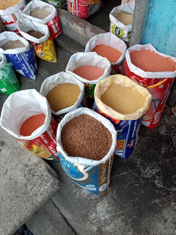 Several open bags filled with various grains and pulses are arranged on a concrete floor. The bags are colorful and patterned, indicating different brands or types of grains. The contents include lentils and other types of seeds or grains. The setting appears to be a marketplace or shop.