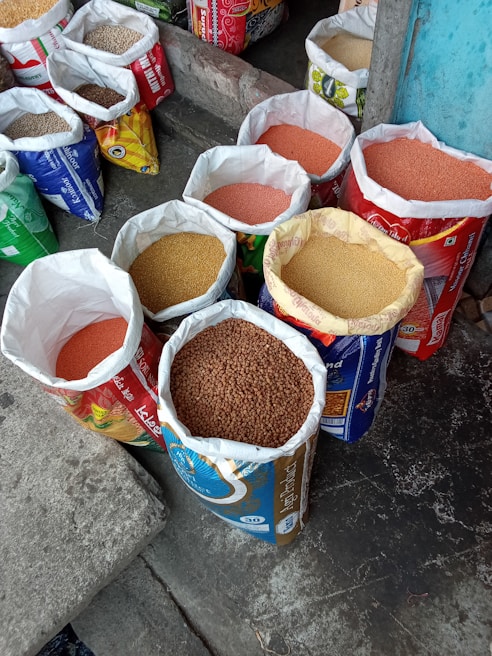 Several open bags filled with various grains and pulses are arranged on a concrete floor. The bags are colorful and patterned, indicating different brands or types of grains. The contents include lentils and other types of seeds or grains. The setting appears to be a marketplace or shop.