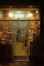 Stacks of books and magazines fill a small, warmly lit bookstore. Shelves are packed tightly, and piles of reading materials line the aisles and floor. A cozy, cluttered atmosphere pervades the space, with fluorescent lights casting a soft glow.