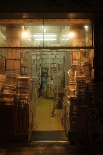 Stacks of books and magazines fill a small, warmly lit bookstore. Shelves are packed tightly, and piles of reading materials line the aisles and floor. A cozy, cluttered atmosphere pervades the space, with fluorescent lights casting a soft glow.