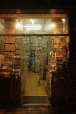 Stacks of books and magazines fill a small, warmly lit bookstore. Shelves are packed tightly, and piles of reading materials line the aisles and floor. A cozy, cluttered atmosphere pervades the space, with fluorescent lights casting a soft glow.