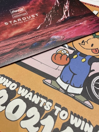 The image features a collection of promotional materials. At the top, there is a Coca-Cola Creations Stardust Limited Edition display with a red outer space theme, including celestial bodies and what appears to be a rocky planetary surface. Below it, there is a colorful cartoon character in blue overalls holding a clock, with part of a car wheel visible in the background. There is also a bold text message suggesting participation in an event or contest with large numbers '2021'.