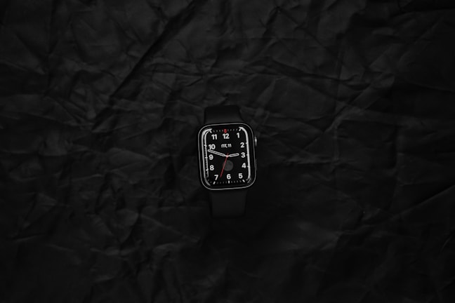 Smartwatch with a minimalist black strap and glowing digital display set on a reflective dark navy background.