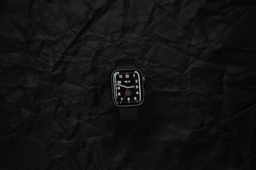 A digital smartwatch with a black band and dark display lies on a textured black surface. The watch face displays white numerals and hour markers with a minimalist design.