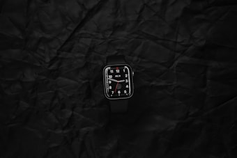 A digital smartwatch with a black band and dark display lies on a textured black surface. The watch face displays white numerals and hour markers with a minimalist design.