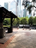 A modern urban park area with a shaded seating area in the foreground, featuring stone pillars and benches. In the background, there are lush green trees and cascading water features creating a tranquil atmosphere. Tall skyscrapers rise above the park, blending nature with the cityscape.