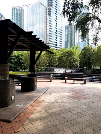 A modern urban park area with a shaded seating area in the foreground, featuring stone pillars and benches. In the background, there are lush green trees and cascading water features creating a tranquil atmosphere. Tall skyscrapers rise above the park, blending nature with the cityscape.
