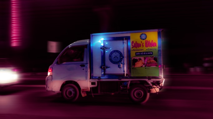 A small truck is driving on a road at night, featuring bright signage on its side advertising a restaurant and catering service. The vehicle is illuminated by purple and blue lights, creating a striking contrast with the dark surroundings.