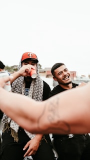 Two people are standing together outdoors, laughing and enjoying what appears to be a social gathering. One person is drinking from a red cup while the other is smiling broadly. Both wear casual attire, with one sporting a cap and a patterned scarf. In the foreground, an arm with a tattoo is visible, possibly capturing a moment on a phone.