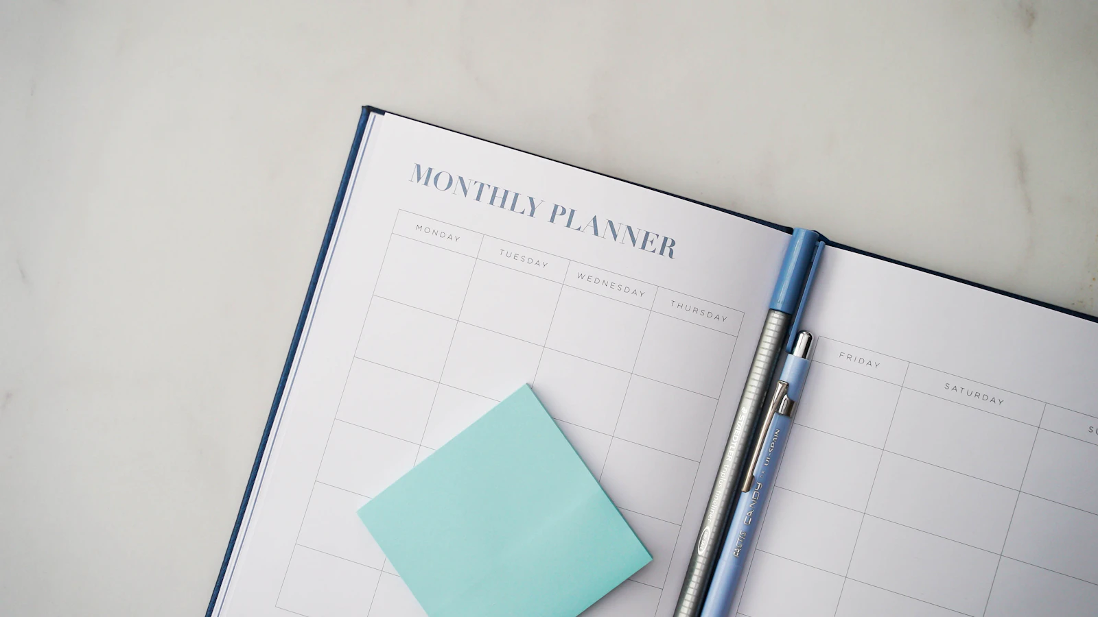 An open monthly planner with a mint sticky note and a pen on a bright desk