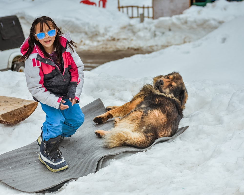 a little girl standing on a snowboard next to a dog