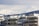 A cityscape featuring modern apartment buildings in the foreground with snow-capped mountains looming in the background. The clouds are thick, casting a muted light over the scene. The buildings have flat roofs with visible antennas and equipment.