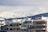 A cityscape featuring modern apartment buildings in the foreground with snow-capped mountains looming in the background. The clouds are thick, casting a muted light over the scene. The buildings have flat roofs with visible antennas and equipment.