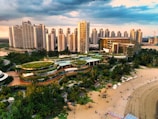 A vibrant urban landscape featuring a series of high-rise buildings with modern architecture. In the foreground, a complex of green-roofed buildings meanders through lush gardens and parkland. A sandy beach lined with people enjoying leisure activities borders the area. The sky is partly cloudy, casting dynamic shadows over the scene.
