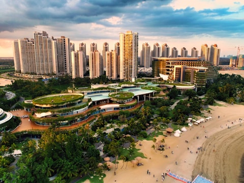 A vibrant urban landscape featuring a series of high-rise buildings with modern architecture. In the foreground, a complex of green-roofed buildings meanders through lush gardens and parkland. A sandy beach lined with people enjoying leisure activities borders the area. The sky is partly cloudy, casting dynamic shadows over the scene.