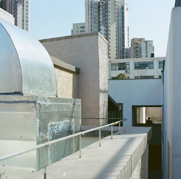 Urban setting with buildings of varying heights, featuring modern architecture. The foreground includes a rooftop area with textured walls and a metallic structure, while high-rise buildings dominate the background.
