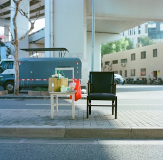 An urban street setting features a black chair positioned next to a white table. On the table, there are various items such as a woven basket with green handles, a red bag, a bottle, and some packaged goods. The background includes a truck, a building, and a leafless tree under an overpass structure.