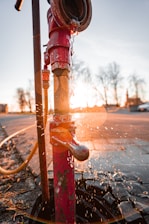 A red fire hydrant with peeling paint is leaking water, creating splashes in the air against the backdrop of a sunset. The sunlight creates a warm glow and lens flare effect around the hydrant.