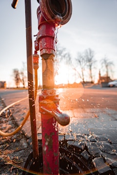 A red fire hydrant with peeling paint is leaking water, creating splashes in the air against the backdrop of a sunset. The sunlight creates a warm glow and lens flare effect around the hydrant.