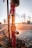 A red fire hydrant with peeling paint is leaking water, creating splashes in the air against the backdrop of a sunset. The sunlight creates a warm glow and lens flare effect around the hydrant.