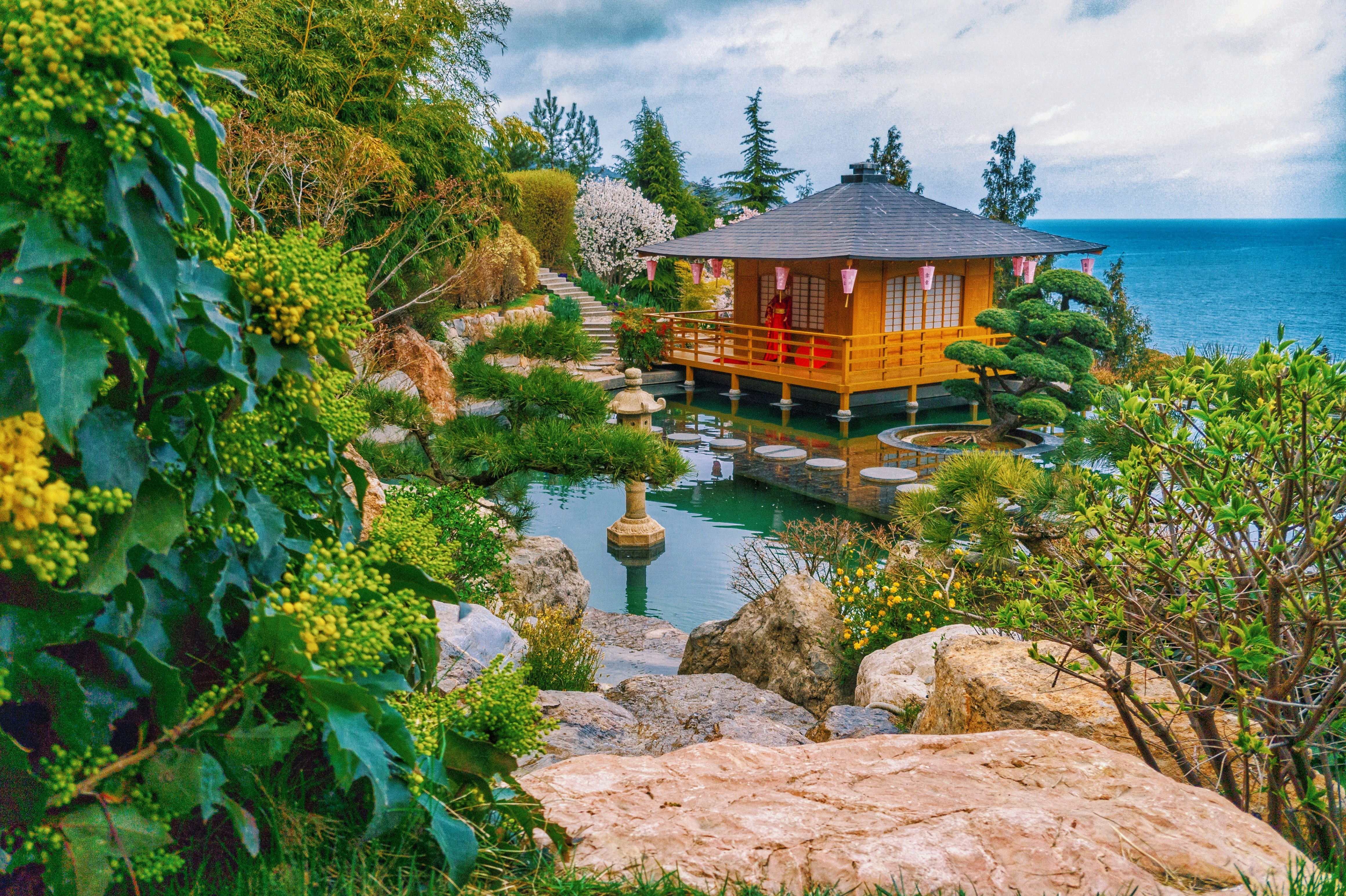 A tranquil Japanese garden featuring a wooden pavilion built over a pond, surrounded by lush greenery, rocks, and stepping stones, with the sea in the background.