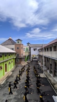A group of people, dressed in black with yellow belts, are practicing martial arts in a lined formation on a narrow street. The buildings on either side are colorful, with bright green and neutral tones, and overhanging roofs. The sky above is clear with some clouds, adding a serene backdrop to the energetic practice.