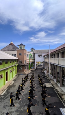 A group of people, dressed in black with yellow belts, are practicing martial arts in a lined formation on a narrow street. The buildings on either side are colorful, with bright green and neutral tones, and overhanging roofs. The sky above is clear with some clouds, adding a serene backdrop to the energetic practice.