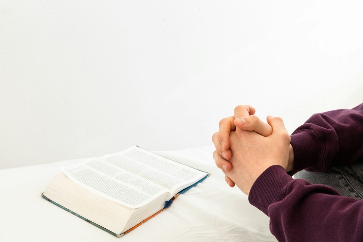 A person sitting at a table with hands resting on an open Bible, praying quietly