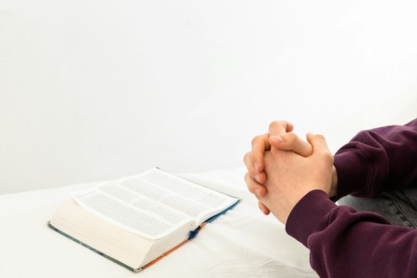 A person sitting at a table with hands resting on an open Bible, praying quietly