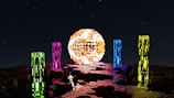 A surreal night scene features a large, glowing spherical object with a mosaic-like surface. Surrounding the sphere are several tall, rectangular pillars, each emitting neon colors: green, pink, blue, and yellow. The ground is a rocky terrain, reflecting the vibrant hues from the pillars and the sphere. The background is a starry night sky, adding a sense of mystique and otherworldliness.