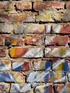 A textured brick wall covered with splashes of colorful paint. The bricks have shades of red, yellow, and orange, and are accented with patches of blue and white paint. The surface of the wall shows signs of wear and age, adding to the rustic and artistic appearance.