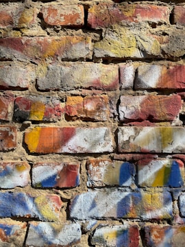 A textured brick wall covered with splashes of colorful paint. The bricks have shades of red, yellow, and orange, and are accented with patches of blue and white paint. The surface of the wall shows signs of wear and age, adding to the rustic and artistic appearance.
