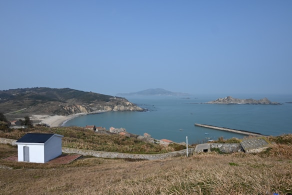 A coastal landscape featuring a small white building on a grassy hillside overlooking a tranquil bay. The bay is bordered by a sandy beach and rocky cliffs in the distance. Several small houses with reddish roofs are visible near the shore. A long pier extends into the calm blue water, and there are rocky islands on the horizon under a clear blue sky.