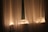 Sheer curtains softly illuminated by warm string lights are draped over a window. The gentle glow from the lights creates a cozy and serene atmosphere. The curtain fabric appears light and airy, allowing a faint view through the window.
