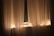 Sheer curtains softly illuminated by warm string lights are draped over a window. The gentle glow from the lights creates a cozy and serene atmosphere. The curtain fabric appears light and airy, allowing a faint view through the window.