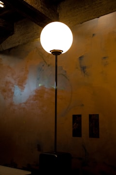 A glowing spherical lamp on a tall stand illuminates a dimly lit room with textured walls. The light casts a warm ambiance, and the wall features abstract patterns in muted tones. Two small rectangular artworks are positioned on the wall.