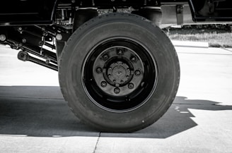 A black and white photo featuring a large truck wheel. The focus is on the massive tire and its intricate hub design, positioned on a concrete surface.