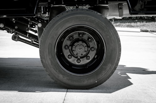 A black and white photo featuring a large truck wheel. The focus is on the massive tire and its intricate hub design, positioned on a concrete surface.