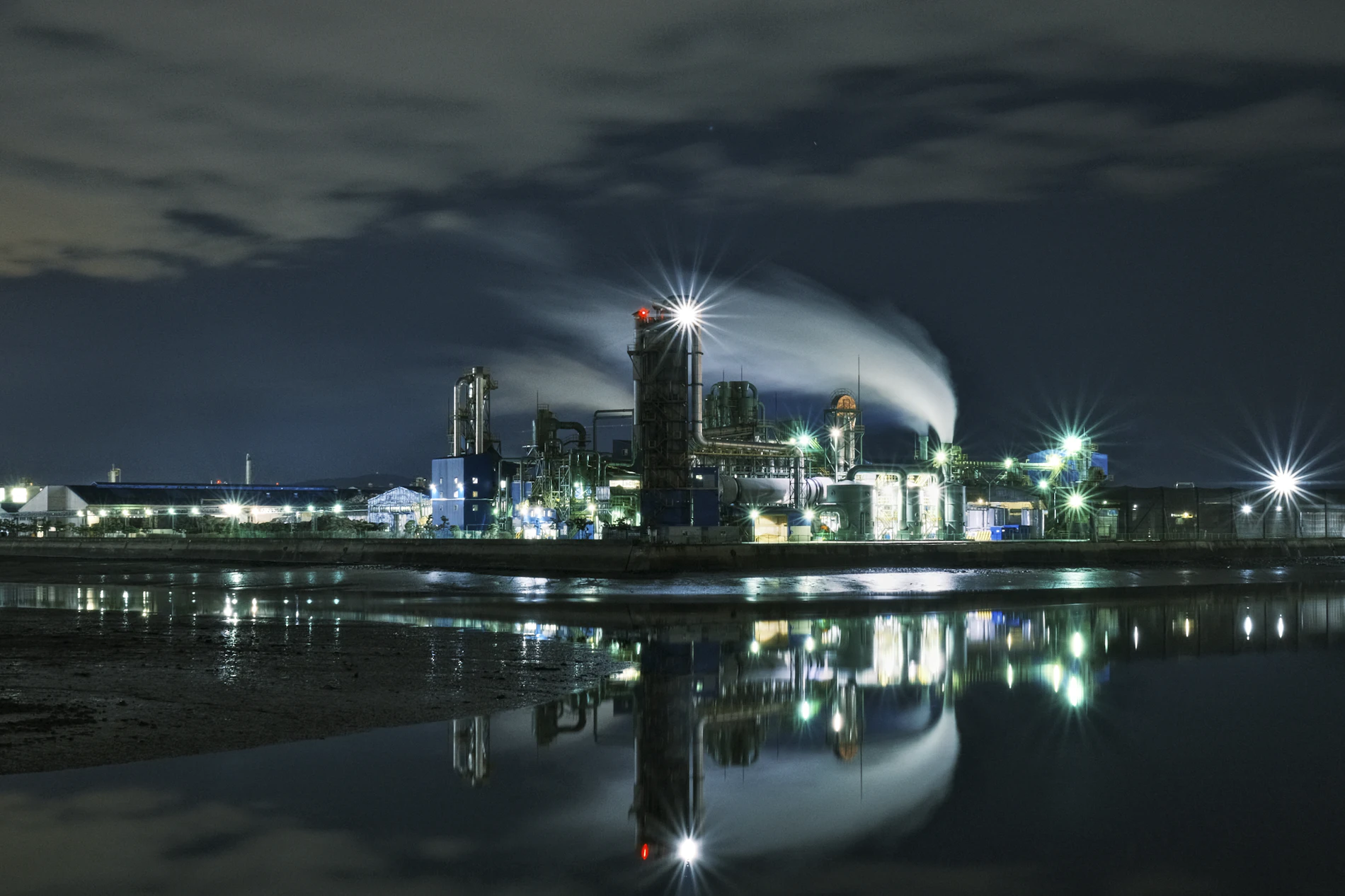 Planta industrial encendida de noche, reflejada sobre el agua.