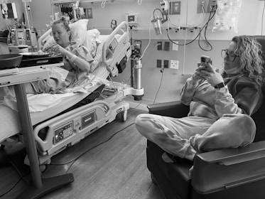 Two individuals are in a hospital room. One person is lying in a hospital bed with medical equipment around, engaging with a tray of food. The other person is sitting in a chair, using a smartphone, dressed in comfortable clothing.